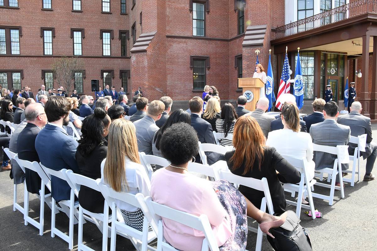 Slavnostní otevření sídla ministerstva vnitřní bezpečnosti USA, řečnice u pultu před cihlovou budovou a řady posluchačů na židlích