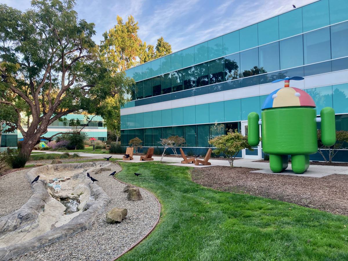 Zelená postava Androidu s vrtulkou před prosklenou budovou Googlu